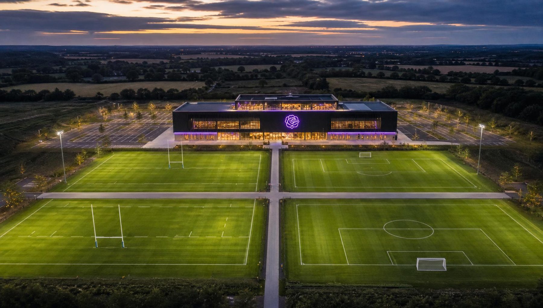 Illustrative dusk aerial render of the proposed campus: one integrated building with a lit purple rose emblem and four full-size pitches arranged in a two-by-two grid