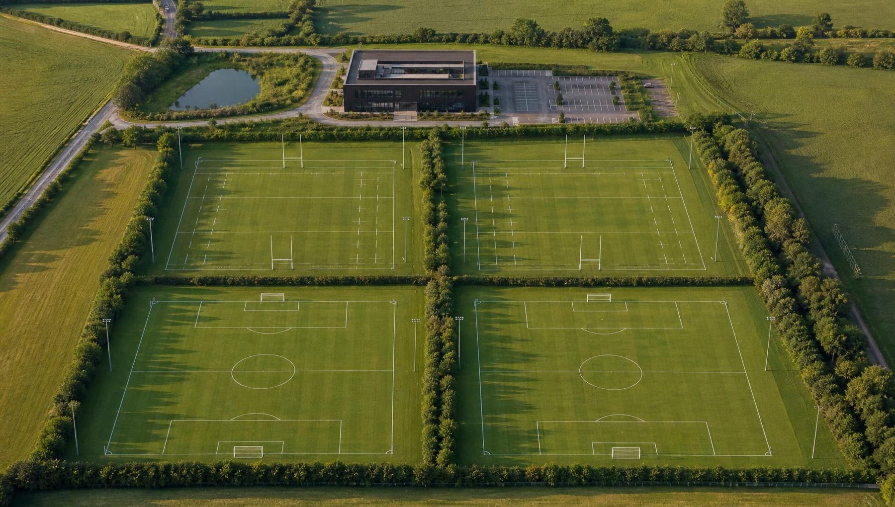 Illustrative aerial masterplan of the proposed campus: one rectilinear building with parking to the east, two rugby pitches with H-posts and two football pitches arranged in a two-by-two grid